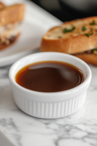 A small white bowl on a white marble cooktop, filled with rich, strained au jus sauce, perfect for dipping the French Dip sandwich, enhancing every bite with deep, savory flavor.