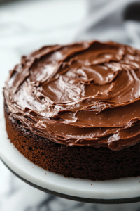 A close-up shot of the cooled chocolate cake on the white marble cooktop as thick, glossy chocolate frosting is spread evenly over the top with an offset spatula, giving the cake a rich and indulgent finish.