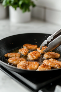 A black skillet on a white marble cooktop, filled with hot oil. Several pieces of almond flour and Parmesan-coated shrimp are frying to a crispy golden brown. A pair of tongs hovers over the pan, ready to flip the shrimp