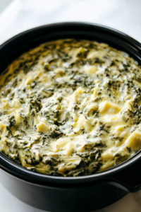 A black slow cooker on a white marble cooktop, with the fully combined and melted spinach artichoke dip. The surface glistens with creamy cheese, and flecks of spinach and artichoke are visible, making it look warm and inviting, ready to be served.
