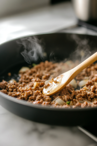 A black skillet on a white marble cooktop sizzling with thinly sliced garlic in olive oil. Lean ground beef is added, browning and crumbling as it cooks. A small spoon of Better Than Bouillon Beef Base is ready to be stirred in for extra flavor.