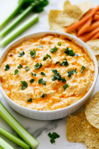 A serving bowl on a white marble cooktop, with Buffalo chicken dip garnished with extra shredded cheese and freshly sliced green onions. The vibrant colors of the toppings contrast beautifully against the creamy dip, making it visually appealing and ready to be enjoyed.