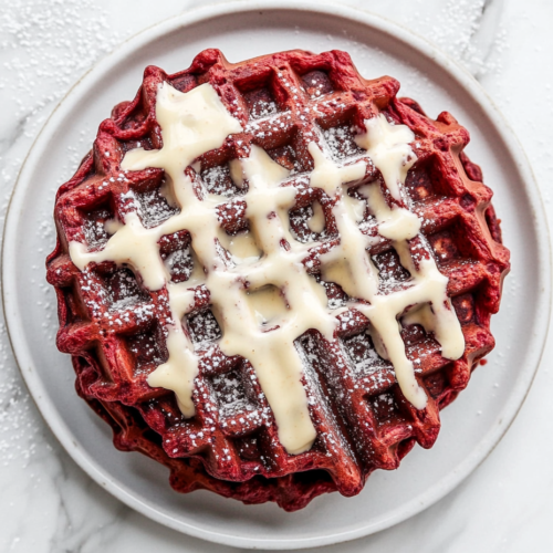 A stack of beet oat waffles or pancakes on a plate, topped with fresh fruit, maple syrup, and a sprinkle of cacao powder, ready to be enjoyed
