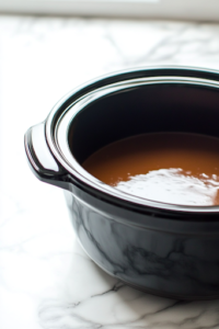 A close-up shot of a black slow cooker sitting on a white marble cooktop, its interior lightly greased with cooking spray. The glistening surface reflects the light, indicating it is prepped and ready for the sweet potatoes to be added.