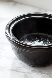 A black slow cooker on a white marble cooktop, lightly coated with cooking spray. The glossy interior is prepped to prevent sticking.