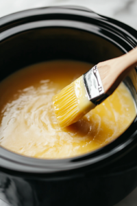 A close-up view of a crockpot being lightly greased with butter using a pastry brush. The butter spreads smoothly over the surface, creating a glistening layer. The crockpot is placed on a white marble cooktop, reflecting a clean and bright kitchen setting.