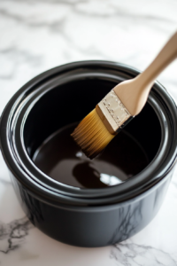 A black slow cooker on the white marble cooktop, its interior being greased with butter using a pastry brush. The shiny surface reflects the light as the butter melts slightly