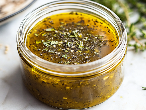 A close-up shot of a sealed airtight container filled with the freshly made keto Greek vinaigrette on the white marble cooktop. A sprinkle of dried oregano sits on top as a garnish, indicating its rich Mediterranean flavor, ready for storage and future use.
