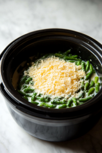 A close-up shot of a black slow cooker on a white marble countertop, filled with frozen green beans, cream of chicken soup, milk, and grated Parmesan cheese before mixing. The ingredients are layered, ready to be stirred into a rich and creamy casserole.