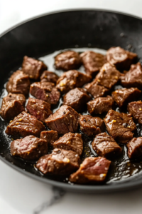 A close-up shot of a black skillet on the white marble cooktop, with hot olive oil shimmering. Freshly diced steak pieces have just been added, sizzling as they begin to sear and develop a rich brown crust.