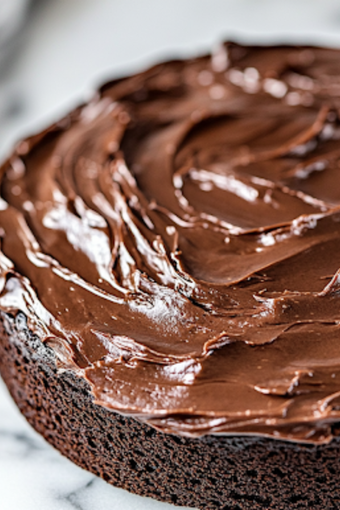 A close-up shot of the cooled chocolate cake on the white marble cooktop as thick, glossy chocolate frosting is spread evenly over the top with an offset spatula, giving the cake a rich and indulgent finish.
