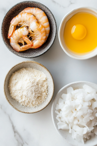 A close-up shot of three small bowls on a wooden countertop, each holding a key ingredient for keto coconut shrimp. One bowl contains an almond flour mixture seasoned with onion powder, garlic powder, salt, and pepper. Another holds two whisked eggs, while the third is filled with a blend of shredded coconut, almond flour, and sweetener. A row of raw jumbo shrimp with tails on is neatly arranged, ready for coating.