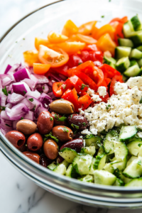 Close-up shot of a glass mixing bowl filled with vibrant Greek salad ingredients including chopped veggies, kalamata olives, and crumbled feta cheese. A wooden spoon rests on the side of the bowl.