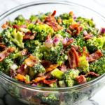 A glass mixing bowl on a white marble cooktop, filled with freshly tossed broccoli bacon salad, covered and ready for refrigeration. The dressing evenly coats the ingredients, with vibrant colors peeking through for a delicious final look.