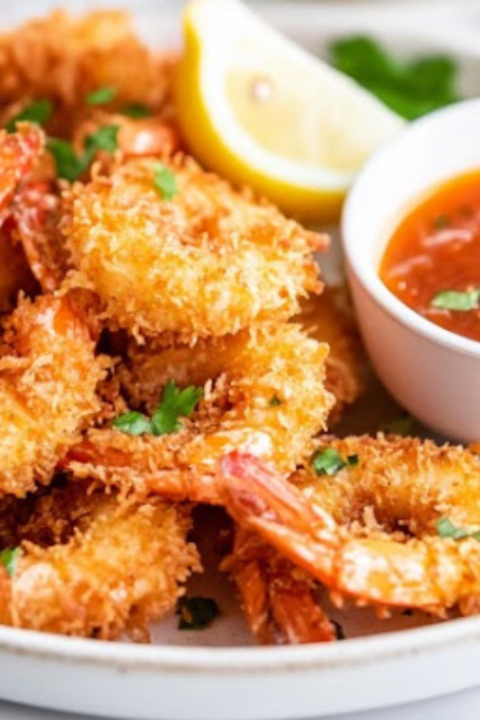 A close-up shot of a plate of crispy keto coconut shrimp, beautifully arranged with a side of tangy dipping sauce. A lemon wedge and fresh herbs garnish the plate, adding a vibrant touch. The shrimp's golden crust glistens under soft lighting, making the dish look restaurant-quality and ready to be enjoyed.