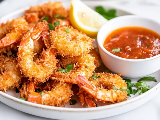 A close-up shot of a plate of crispy keto coconut shrimp, beautifully arranged with a side of tangy dipping sauce. A lemon wedge and fresh herbs garnish the plate, adding a vibrant touch. The shrimp's golden crust glistens under soft lighting, making the dish look restaurant-quality and ready to be enjoyed.