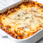 A freshly baked keto eggplant lasagna in a white ceramic baking dish on a white marble cooktop. The top is covered with bubbling, golden-brown mozzarella and Parmesan cheese, resting for 10 minutes before slicing.
