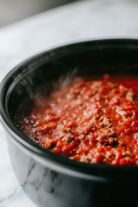 A close-up shot of a black slow cooker on the white marble cooktop with a layer of meat sauce spread evenly across the bottom. The deep red sauce glistens under the light, forming the flavorful base for the crockpot lasagna.