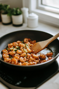 Close-up shot of the skillet on the white marble cooktop, where the shrimp are now fully pink and the honey-lime sauce is thickening into a glossy glaze. A wooden spoon stirs the shrimp to coat them evenly