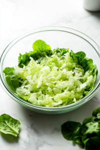 A large glass serving bowl on a white marble cooktop, filled with freshly washed and shredded lettuce, evenly spread at the bottom. A few crisp lettuce leaves sit beside the bowl, adding freshness.