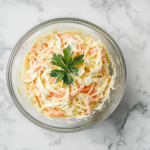 A close-up shot of the completed Keto Coleslaw in a large glass mixing bowl on a white marble cooktop. The creamy dressing has evenly coated the vibrant shredded vegetables, with a sprig of fresh parsley garnishing the top, ready to be served.