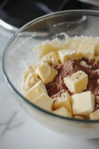 A glass mixing bowl on a white marble cooktop contains flour, ground hazelnuts, baking powder, salt, cinnamon, and sugar, being mixed together. Cold cubes of vegan butter and cold water are added to form dough.