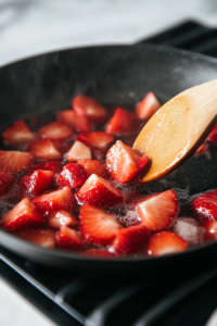A saucepan on a stovetop with fresh strawberries, sugar, and lemon juice simmering into a soft, vibrant red sauce.