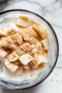 Close-up shot of a glass food processor on the white marble cooktop, filled with all-purpose flour, raw cane sugar, cubed cold vegan butter, dairy-free milk, and sea salt. The mixture is being pulsed into a crumbly dough.