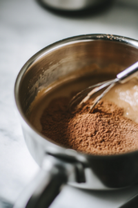 Close-up shot of a shiny stainless steel saucepan on the white marble cooktop, with soy milk, granulated sugar, and cornstarch being whisked together. The mixture slowly thickens over low-medium heat, with steam rising as a hand holds the whisk steadily.
