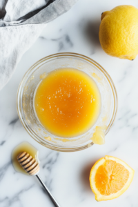 Close-up shot of a small glass bowl on a white marble countertop, with orange juice, lemon juice, honey, vanilla extract, and citrus zest being whisked together into a smooth dressing.