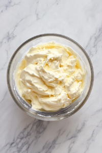 Cold heavy cream is being whipped into medium peaks in a mixing bowl on the white marble cooktop with a hand mixer. Powdered sugar and vanilla sit beside the bowl