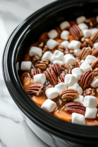A close-up shot of the black slow cooker on the white marble cooktop, now uncovered, revealing the tender, caramelized sweet potatoes. A generous handful of mini marshmallows and whole pecans have been sprinkled over the top, ready to melt and add texture.