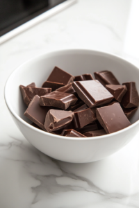 A microwave-safe bowl on the white marble cooktop, filled with chunks of chocolate milk or dark, melting in 20-second bursts. The chocolate begins to soften and melt smoothly.