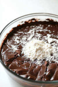 Close-up shot of the same glass mixing bowl on the white marble cooktop, as the cooled melted chocolate mixture is being carefully folded into the whipped egg mixture with a spatula. The batter looks rich and velvety