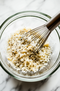 A glass mixing bowl on a white marble cooktop, filled with almond flour, grated Parmesan cheese, kosher salt, and black pepper. A stainless steel whisk is blending the ingredients into a fine, even mixture.