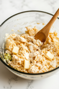 A large glass mixing bowl on the white marble cooktop is filled with drained chunk chicken, cooked rice, tender broccoli, cubed processed cheese, cream of mushroom soup, cream of chicken soup, milk, cubed butter, and chopped onion. A wooden spoon stirs the thick and creamy mixture, combining all the flavors.