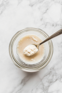 A small glass bowl on a white marble cooktop, with cornstarch being mixed with water. A spoon stirs the mixture until it forms a smooth, thick slurry, which will help thicken the sauce later.