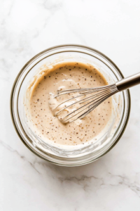 A close-up shot of a glass mixing bowl on the white marble cooktop, filled with condensed cream of mushroom soup, water, garlic powder, salt, and black pepper. A whisk is inside the bowl, blending the ingredients into a smooth, flavorful sauce.