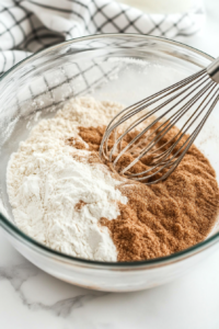 A mixing bowl on a wooden countertop, filled with flour, brown sugar, white sugar, cinnamon, nutmeg, and salt. A whisk is in the bowl, evenly blending the ingredients for the topping.