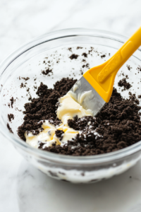A close-up shot of a glass mixing bowl on a white marble countertop, where Oreo crumbs and melted butter are being combined with a spatula, creating a moist crumb mixture.