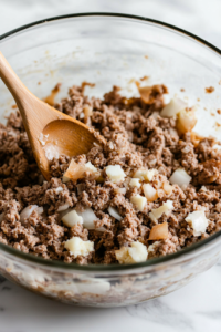 A close-up of a large clear glass mixing bowl on a white marble cooktop, filled with ground beef, minced onion, dried stuffing mix, milk, eggs, salt, and pepper. A wooden spoon is stirring the ingredients, combining them into a smooth, well-mixed meatloaf mixture.