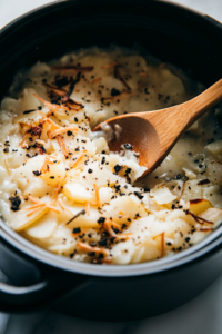A black crockpot on a white marble cooktop filled with thawed hash browns, diced potatoes, chopped onions, and black pepper. A wooden spoon stirs the mixture with chicken broth and cream of chicken soup, combining all the ingredients for a rich, flavorful base.