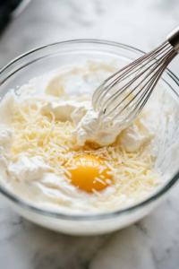 A glass mixing bowl on a white marble cooktop holding creamy ricotta cheese, a cracked egg, and shredded mozzarella. A metal whisk blends the ingredients into a smooth, cheesy mixture, ready for layering.