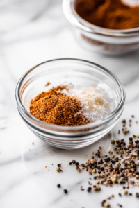 A close-up of a small glass bowl on a white marble cooktop, filled with a well-balanced seasoning mix of brown sugar, salt, pepper, garlic powder, onion powder, and smoked paprika. The blend is ready to be applied to enhance the chicken’s flavor.