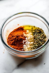 A close-up of a small glass bowl on a white marble cooktop, filled with a vibrant seasoning blend of garlic powder, onion powder, paprika, salt, and pepper. Olive oil is drizzled in, and a spoon stirs everything together, creating a rich, aromatic rub for the turkey.