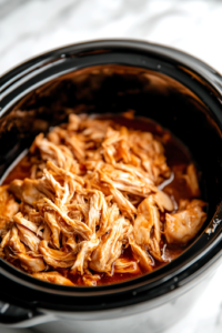 A black slow cooker on a white marble cooktop, with freshly shredded chicken thoroughly mixed into the rich barbecue sauce. The sauce thickens as it coats every strand of chicken, creating a juicy and flavorful filling.