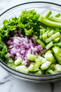 The same glass serving bowl on a white marble cooktop, now layered with diced red onions and sliced celery over the lettuce. The vibrant purple and green ingredients add contrast and texture.