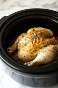 A close-up of a black slow cooker on a white marble cooktop, as the seasoned whole chicken is carefully placed breast-side up inside. The chicken is positioned for even cooking and maximum tenderness.