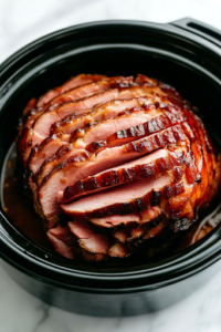A close-up shot of an 8 lb spiral-cut ham placed inside the black crockpot on the white marble cooktop, flat side down. The neatly sliced layers of pinkish-brown meat are arranged in a spiral pattern, ready to absorb the flavors during slow cooking.