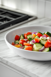 Final plated shot of a beautifully arranged Greek salad in a rustic ceramic bowl, garnished with extra feta cheese and a sprinkle of oregano. A fork rests on the side, and a glass of white wine sits in the background.
